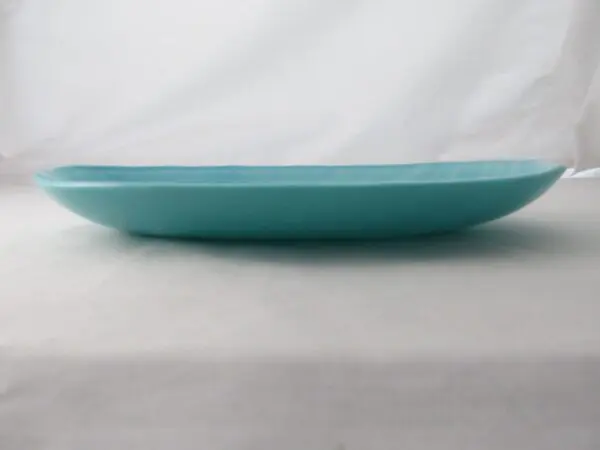 An aquamarine frost oval serving dish, viewed from the side, is placed on a white surface with a white background.