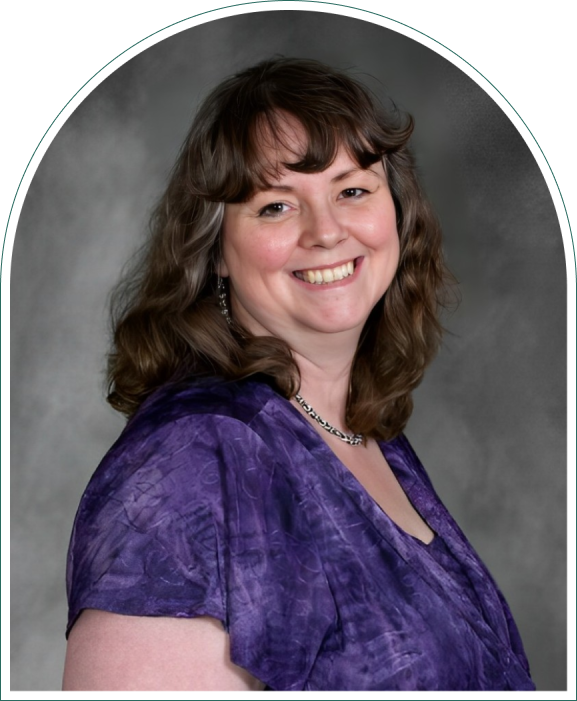 A person with long, wavy brown hair is smiling, wearing a purple blouse and a necklace, in front of a gray backdrop.